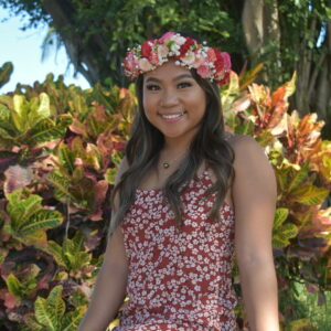 Rochelle sitting on a rock wall, wearing a red and write floral dress and haku lei. Rochelle has wavy brown hair.